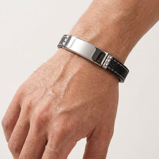 Aramat Jewels zwarte leren herenarmband met graveerbare zilverkleurige 316L RVS plaat en wit stiksel om een pols.