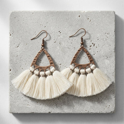 Aramat Jewels boho oorhangers met creme kwastjes en gehamerd bronskleurig metaal op een grijze stenen ondergrond.