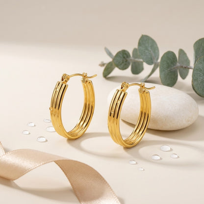 Aramat Jewels gold oval hoop earrings with ribbed texture and small dot details, 26mm. Styled with eucalyptus leaves and a smooth stone.