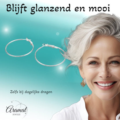 Aramat Jewels silver-colored metal hoop earrings, 40mm diameter, with a clasp closure. A smiling woman with grey hair wears a pair.