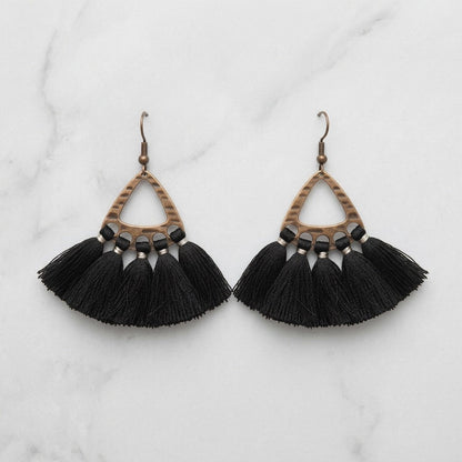 Aramat Jewels boho oorhangers met zwarte kwastjes en een bronskleurig metalen frame op een marmeren achtergrond.